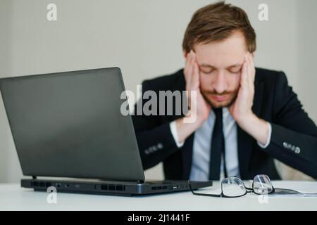 Turbato giovane uomo d'affari in vestito che lavora su un computer portatile e massaggiante testa sentire dolore emicrania, sottolineato fuori. Foto Stock