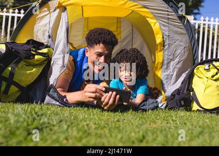 Felice padre e figlio ispanico che guarda il video sullo smartphone mentre si trova all'interno della tenda sul cortile Foto Stock