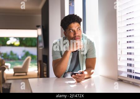 Il pensieroso uomo ispanico si guarda via mentre si appoggia con lo smartphone sul banco della cucina a casa Foto Stock