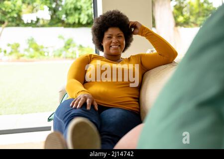 Ritratto a tutta lunghezza di sorridente afroamericana, donna adulta seduta sul divano a casa Foto Stock