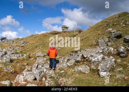 Un camminatore in una giacca rossa brillante che cammina su un sentiero accidentato fino alla scogliera di Cheesewring su Stowes Hill a Bodmin Moor in Cornovaglia. Foto Stock