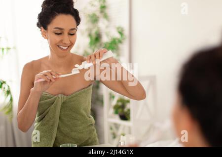 Giovane donna con un bel sorriso che indossa un asciugamano dopo il bagno, applicando dentifricio sul pennello di fronte allo specchio a casa Foto Stock