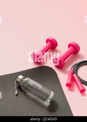 Elegante palestra grigia e rosa. Vista prospettica di tappeto sportivo grigio, bottiglia d'acqua, manubri rosa e corda da salto su sfondo rosa. Set per pilates, fitness con spazio di copia Foto Stock