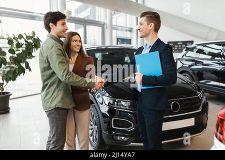 Felice giovane coppia caucasica che acquista auto nuova, scuotendo le mani con sicuro venditore in concessionaria Foto Stock
