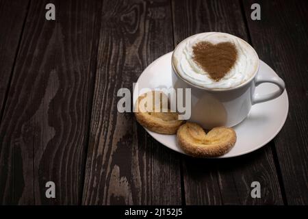 caffè o tè caldo con panna montata con biscotti a forma di cuore e decorazioni su sfondo di legno scuro con spazio copia Foto Stock