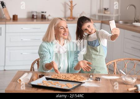 La donna matura e la sua nipote che prendono selfie mentre preparano i biscotti in cucina Foto Stock