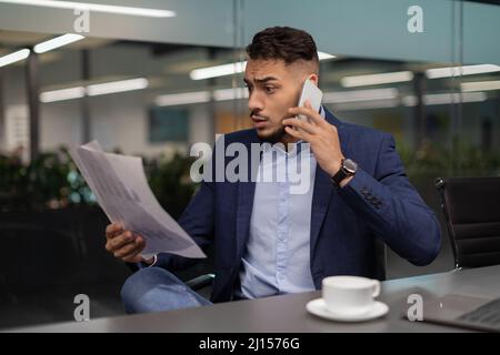 Un uomo d'affari arabo arrabbiato che legge i documenti e ha una conversazione telefonica Foto Stock