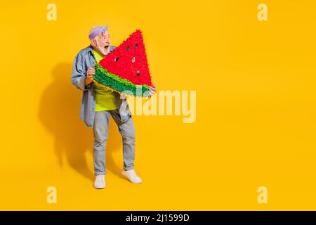 Foto di divertente impressa ragazzo pensionato vestito jeans camicia mordente fetta di cocomero spazio vuoto isolato colore giallo sfondo Foto Stock