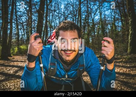 Giovane uomo sta facendo trekking nel bosco. Il bellissimo uomo ha una giacca blu, uno zaino rosso e bastoncini da trekking. Foto Stock