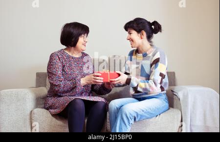 Una giovane donna dona a una donna anziana un regalo in scatola rossa sul suo divano a casa Foto Stock