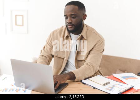 Il giovane freelance africano concentrato si siede al tavolo e guarda al notebook. Serio contabile nero che lavora in linea con i dati del calcolatore Foto Stock