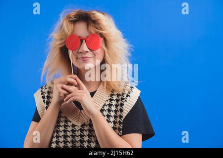 Concetto di cabina fotografica. Giovane allegra donna caucasica sorridente nel suo 20s utilizzando falsi occhiali rossi per coprire gli occhi. Foto studio con sfondo blu. Foto di alta qualità