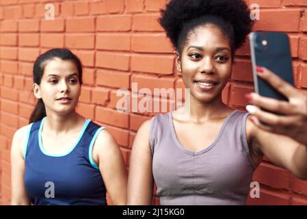 Immagine di donne atletiche giovani sorprendenti che prendono un selfie contro un muro di mattoni. Le donne nere sorridenti trascorrono il tempo insieme dopo l'allenamento sportivo. Foto Stock