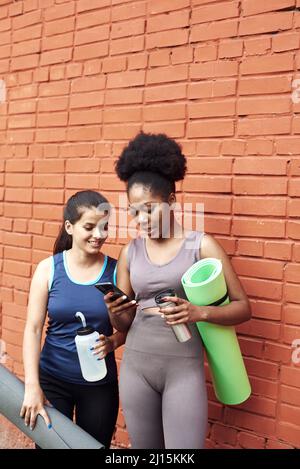 Immagine di donne atletiche giovani sorprendenti di fronte a un muro di mattoni, guardando nel telefono. Due amici di sesso femminile ridono al post sui social media. Foto Stock