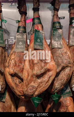 Pamplona, Spagna - 20 giugno 2021: Gambe bone-in intere di prosciutto spagnolo serrano iberico appese in mostra ad un macellaio nella città vecchia o casco Viejo Foto Stock