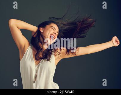 Ha un'esplosione ovunque vada. Studio girato di una giovane donna spensierata gridando con le braccia sollevate e i capelli soffiano su uno sfondo scuro. Foto Stock