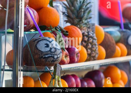 primo piano di una noce di cocco con occhi dipinti e un sorriso, arance, melograni e altri frutti a vista di un bar con succo appena spremuto Foto Stock