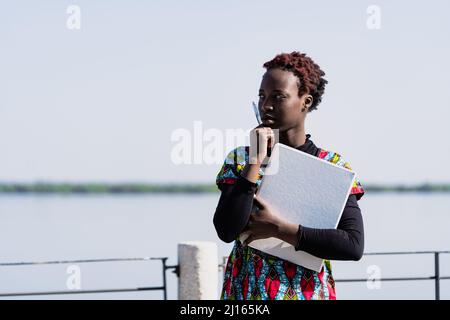 Giovane studentessa africana pensiva che abbraccia il suo laptop e tiene in mano una matita, in piedi sul fiume pensando agli esami del giorno successivo; prova a. Foto Stock