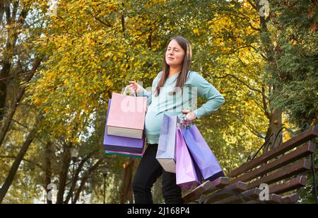 Mentre camminando lungo la strada, la donna incinta comincia ad avere contrazioni uterine. Madre con i pacchetti nelle sue mani dopo lo shopping in piedi alla panca, tenendo la sua vita. Foto Stock