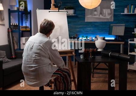 Donna anziana che si sente ispirata guardando creativo matita disegno di vaso su cavalletto seduta in studio d'arte professionale. Artista anziano in serata finendo ombreggiando disegno bianco e nero. Foto Stock