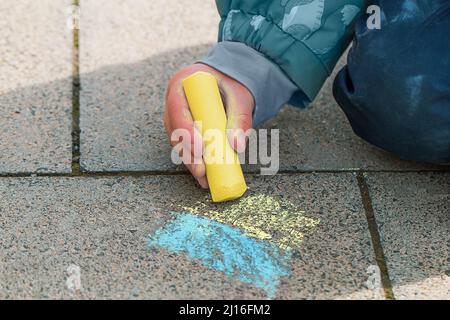 Il bambino disegna la bandiera Ucraina sul marciapiede. La mano di un bambino tiene un gesso giallo. Banner di supporto per l'Ucraina. Foto Stock