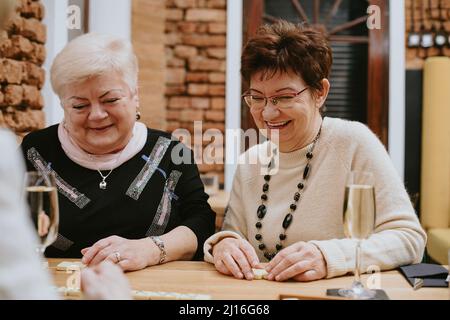 Due sorridenti donne anziane dai capelli scuri e dai capelli discrete si siedono al tavolo di legno con bicchieri di champagne che giocano a domino. Foto Stock