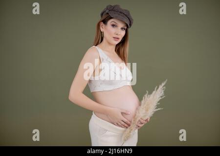 Ritratto di giovane adorabile donna incinta con lunghi capelli scuri che indossa cappello marrone che tiene il ventre e pampas erba secca. Foto Stock