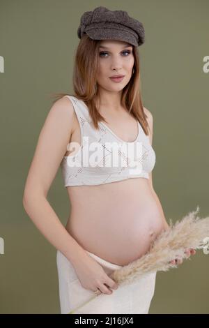 Ritratto di giovane donna incinta impeccabile con lunghi capelli scuri che indossa cappello marrone che tiene il ventre e pampas erba secca. Foto Stock