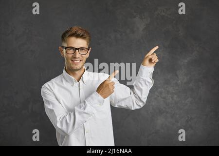 L'uomo amichevole con espressione sorridente punta le dita dell'indice verso il lato su sfondo grigio. Foto Stock