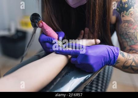 Primo piano della macchina tatuaggio. Un maestro di tatuaggio femminile in una maschera e guanti crea un modello sulla pelle di una donna. Foto Stock