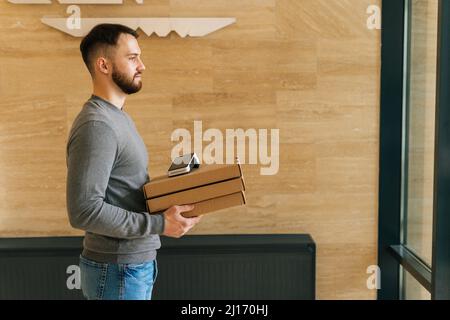 Vista laterale del corriere bearded maschio che tiene le scatole della pizza e del terminale di POS di pagamento contactless, in piedi nella sala d'ingresso dell'edificio dell'appartamento o dell'ufficio Foto Stock