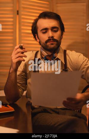 uomo con baffi guardando la carta e tenendo sigaro vicino macchina da scrivere retrò sulla scrivania Foto Stock