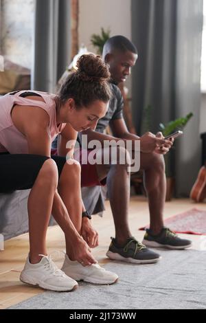 Colpo verticale della giovane donna caucasica che aggraffa le sue sneakers, il suo ragazzo nero che guarda qualcosa sullo smartphone mentre la aspetta Foto Stock