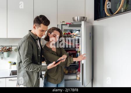 Giovane uomo con donna che controlla la lista sul tablet PC da frigorifero in cucina Foto Stock