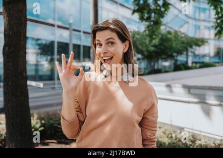 Donna allegra gesturante segno ok con mano in piedi al parco ufficio Foto Stock