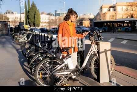 Giovane donna che sblocca la bicicletta elettrica al parcheggio della città Foto Stock