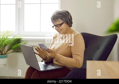 Donna anziana felice seduta in poltrona, utilizzando laptop e smartphone, messaggi di testo e sorridente Foto Stock