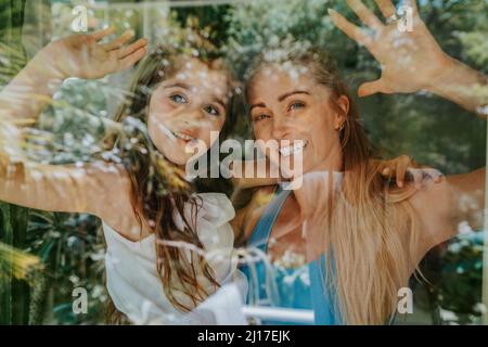 Madre e figlia sorridenti che ondeggiavano attraverso la finestra di casa Foto Stock