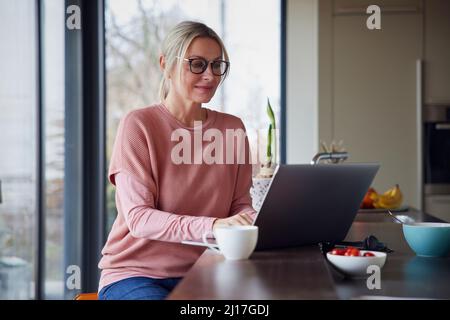 Donna bionda che usa un computer portatile in cucina a casa Foto Stock
