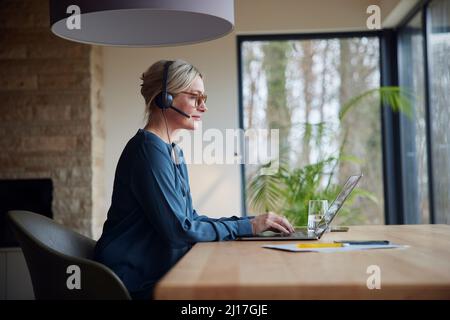 Donna bionda che indossa le cuffie utilizzando un computer portatile seduto al tavolo Foto Stock