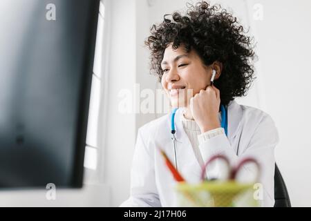 Medico sorridente seduto con la mano sul mento ascoltando le cuffie intrauricolari alla clinica Foto Stock