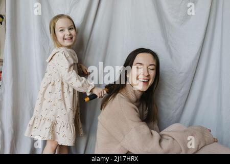 Ragazza bionda sorridente pettinando i capelli della madre a casa Foto Stock