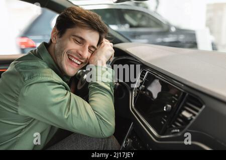 Felice giovane uomo seduto sul sedile del conducente, abbracciato volante della sua nuova vettura, occhi chiusi e sorridenti, spazio copia Foto Stock