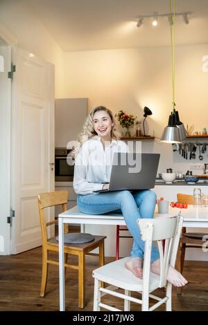 Allegro freelance con computer portatile seduto sul tavolo in cucina a casa Foto Stock