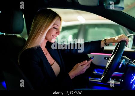 Donna bionda sorridente con smartphone seduto in auto Foto Stock