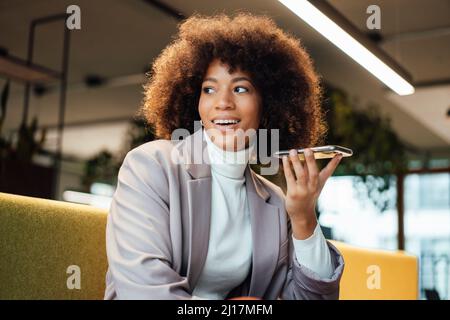 Donna d'affari sorridente che invia messaggi vocali tramite smartphone in ufficio Foto Stock