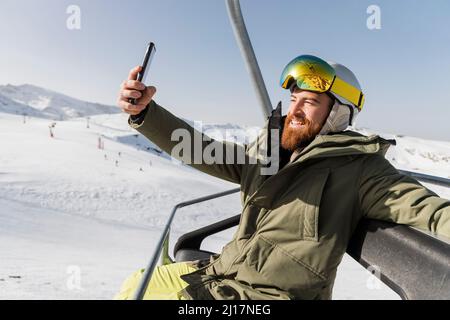 Sorridente giovane uomo che prende selfie attraverso il telefono cellulare seduto sullo skilift Foto Stock