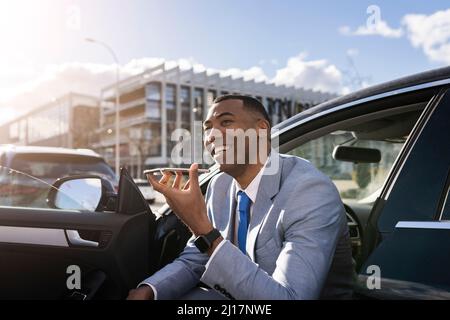 Un uomo d'affari allegro che invia la segreteria telefonica tramite telefono cellulare seduto in auto il giorno di sole Foto Stock
