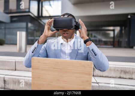 Uomo d'affari che indossa simulatore di realtà virtuale seduto con un laptop sui gradini Foto Stock