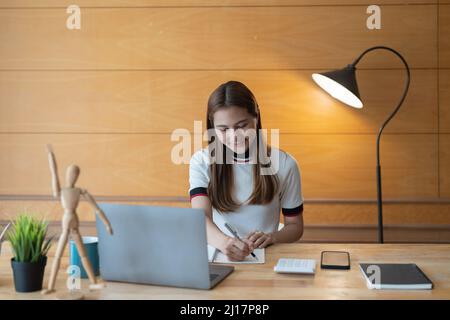 Giovane ragioniere asiatico prendendo nota su blocco note e utilizzando un computer portatile, calcolatrice per finanziario. Foto Stock